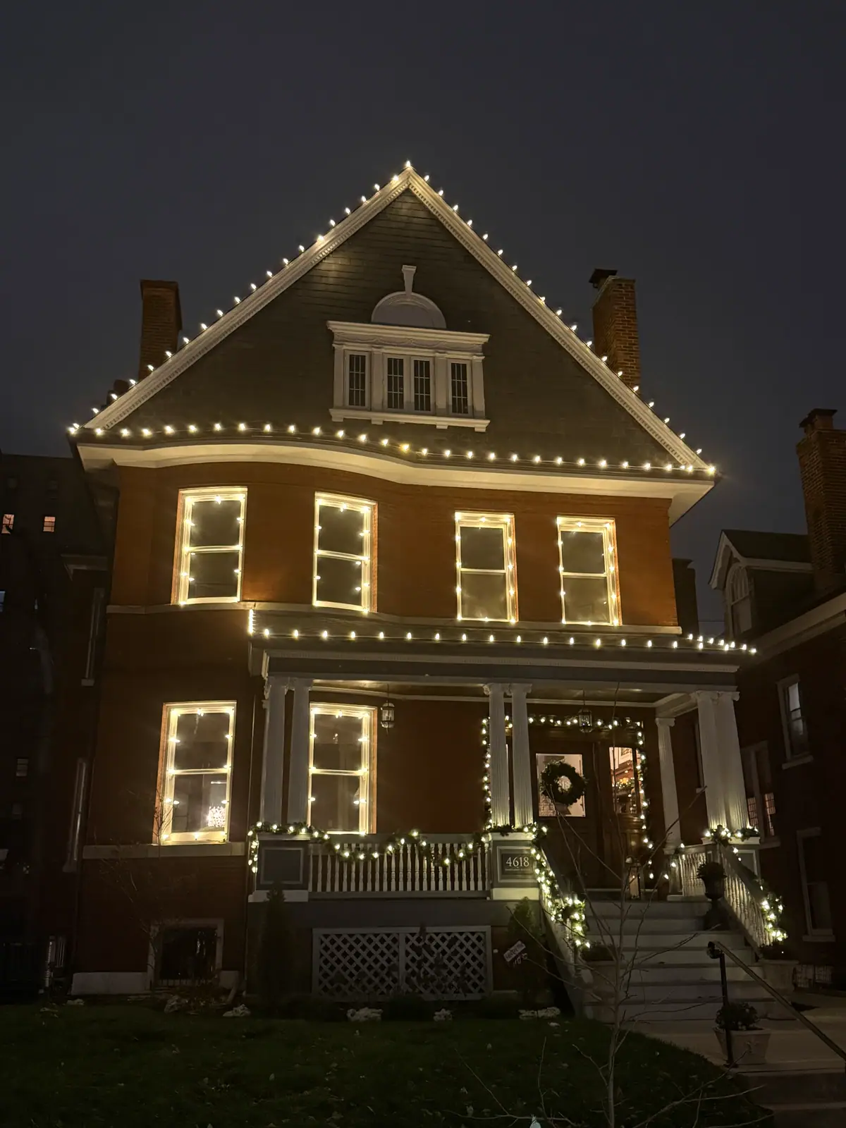 A historic brick two-story outlined in warm white lights, every window glowing and garland down the porch