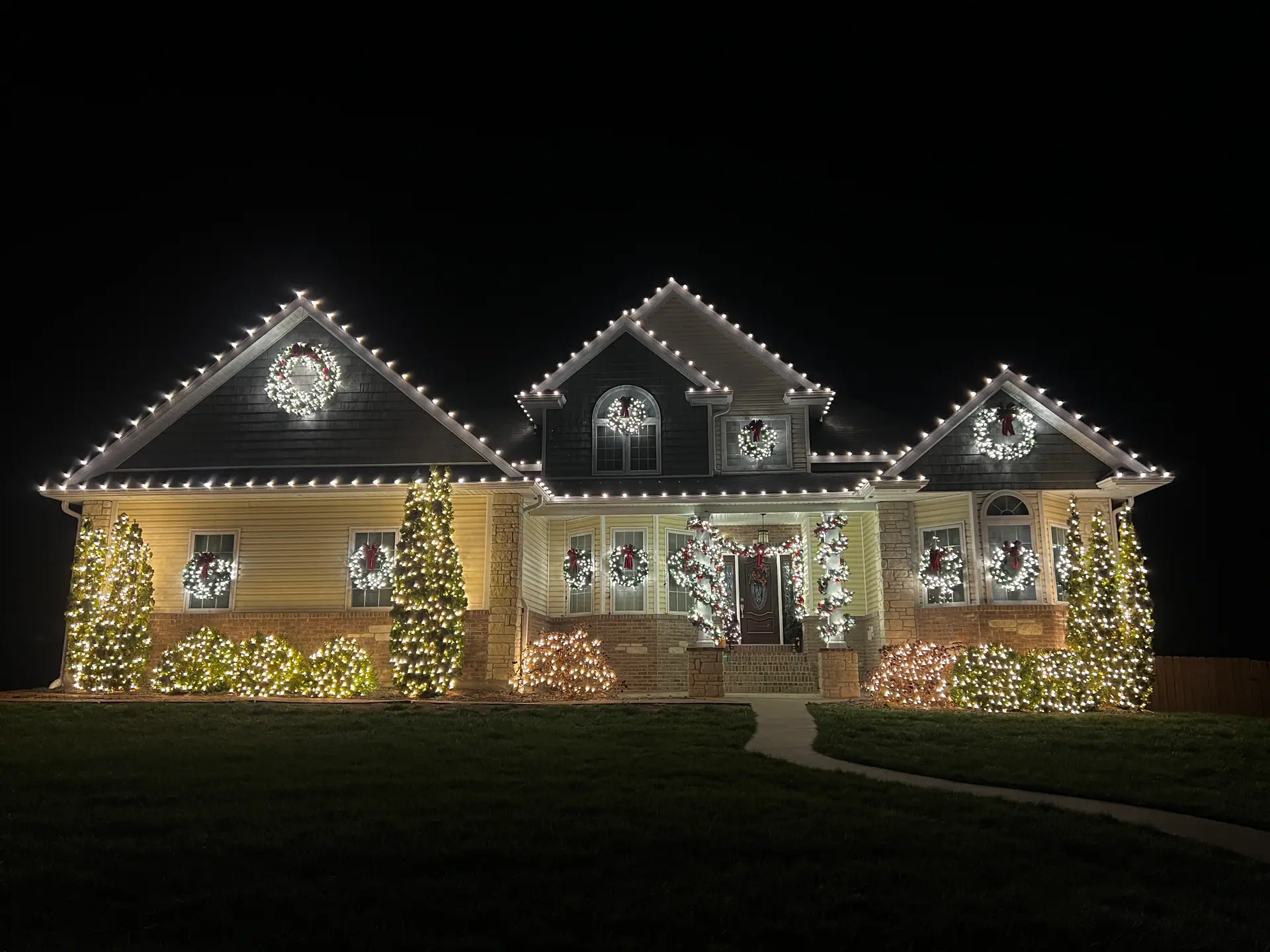 A two-story home lit for Christmas with warm white rooflines, a lit wreath in every window, and shrubs wrapped front to back